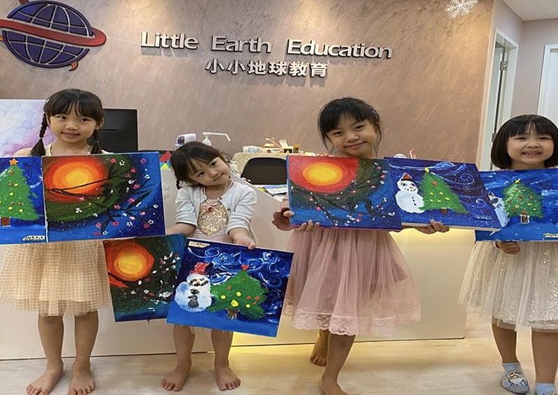 Four girls stand at the Little Earth reception desk holding up their paintings of Christmas trees, snowmen and a glowing sun
