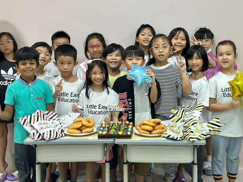 A big group of children, many in Little Earth T-shirts, crowd round two tables of doughnuts and a birthday cake, smiling and making peace signs