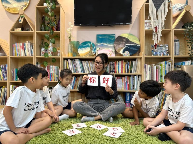 A Little Earth teacher holds up the Chinese characters 你好 on flashcards while five children in Little Earth T-shirts sit on the carpet and read along