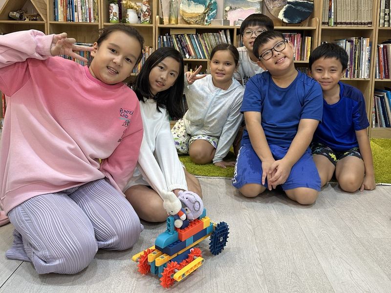 Six children kneel on the floor by the bookshelves, smiling and waving, with the robot they built from a construction kit in front of them